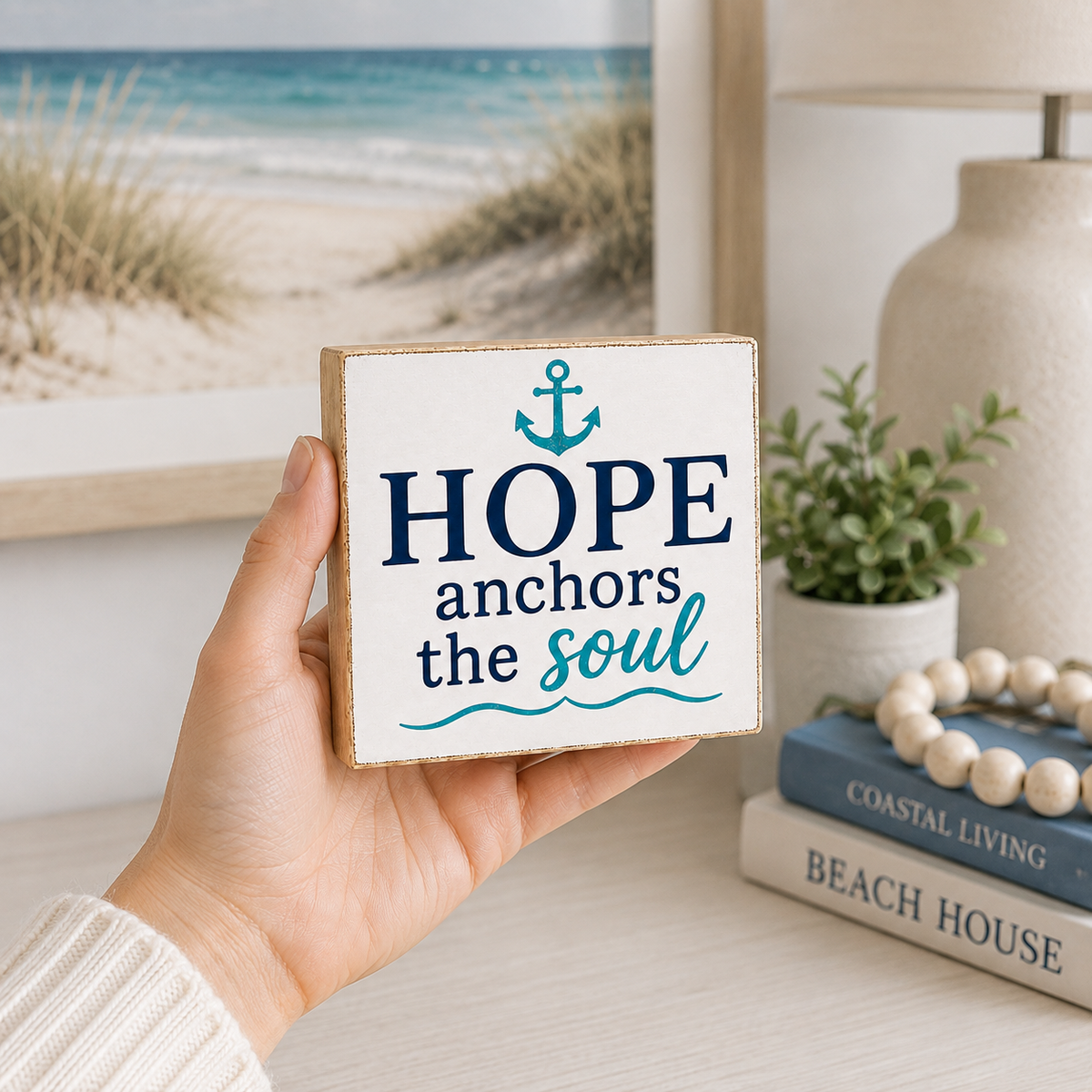 A hand holds a square wooden sign with the phrase 'HOPE anchors the soul' in navy and teal lettering, with a teal anchor graphic at the top. The background shows a softly blurred beach-themed room setting with a framed beach print, a lamp, books, and a small plant.