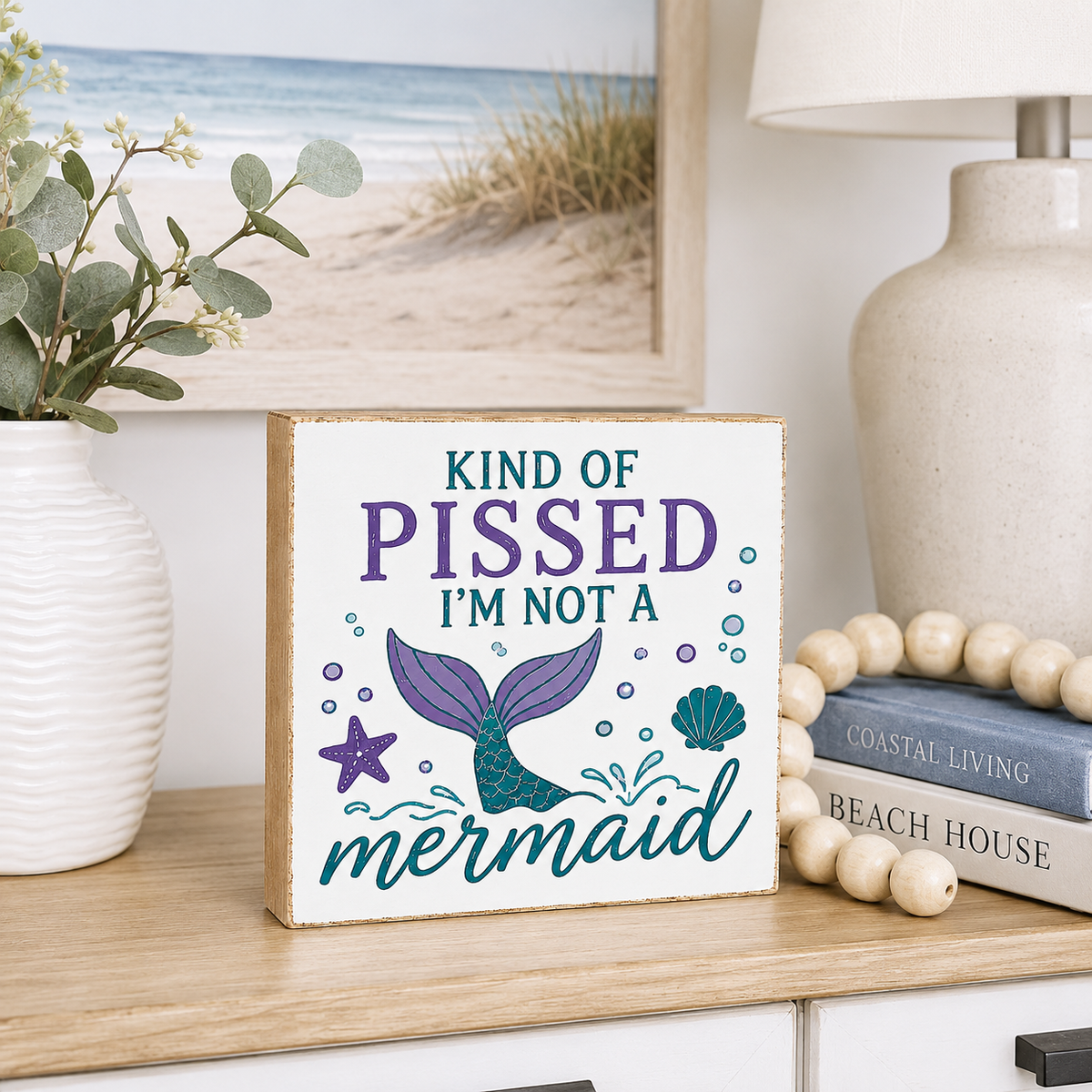 A square decorative sign with a mermaid tail illustration and the text 'KIND OF PISSED I'M NOT A mermaid' in purple and teal lettering, styled on a wooden shelf beside a vase, books, and a lamp in a beach-themed room setting.