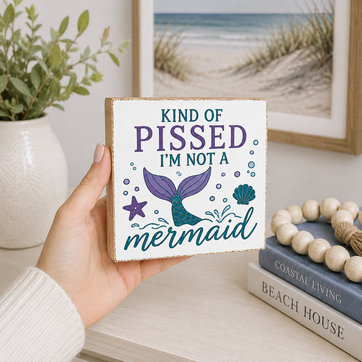 A hand holds a square wooden sign with a mermaid design and the text 'KIND OF PISSED I'M NOT A mermaid' in a beach-themed room setting.