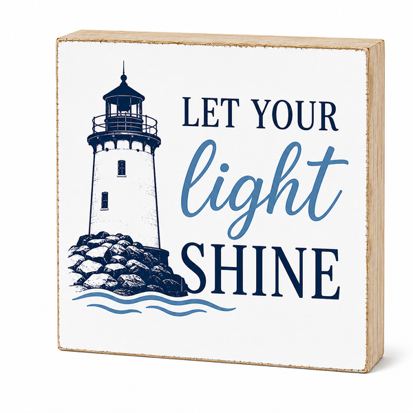 A square decorative wooden sign with a light natural wood border, featuring a navy blue lighthouse illustration and the phrase 'LET YOUR light SHINE' on a white front panel, shown against a light gray background.