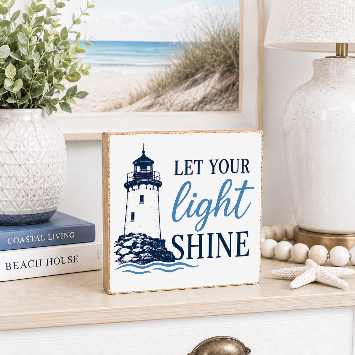 A square decorative sign with a lighthouse illustration and the phrase “LET YOUR light SHINE” in navy and blue lettering, styled on a tabletop with a vase, books, lamp, beads, and a starfish nearby.