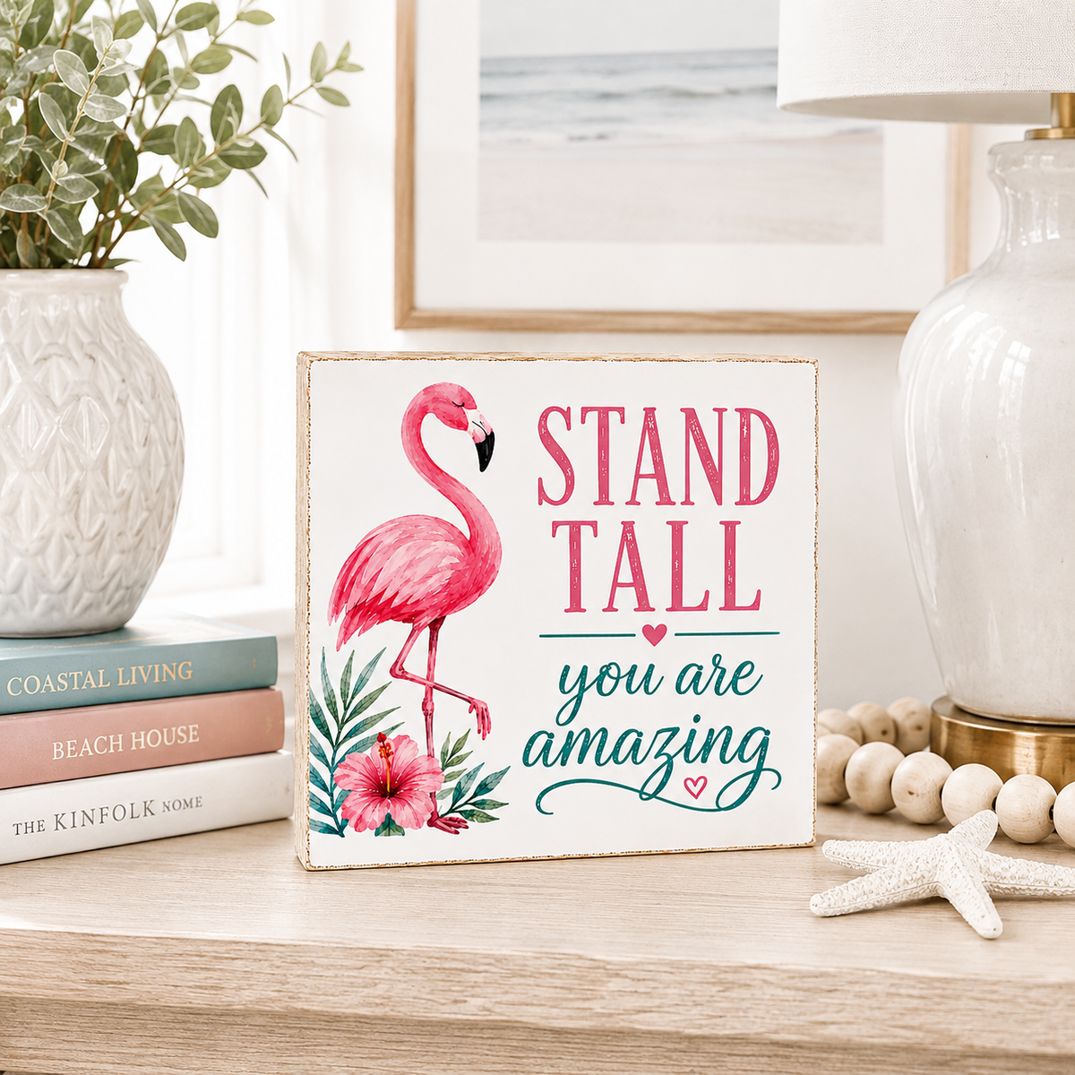 A square decorative sign with a rustic white background featuring a pink flamingo illustration and the text 'STAND TALL you are amazing' in pink and teal lettering. The sign is styled on a light wood table next to a white vase with greenery, a stack of books, a white lamp, and a starfish.