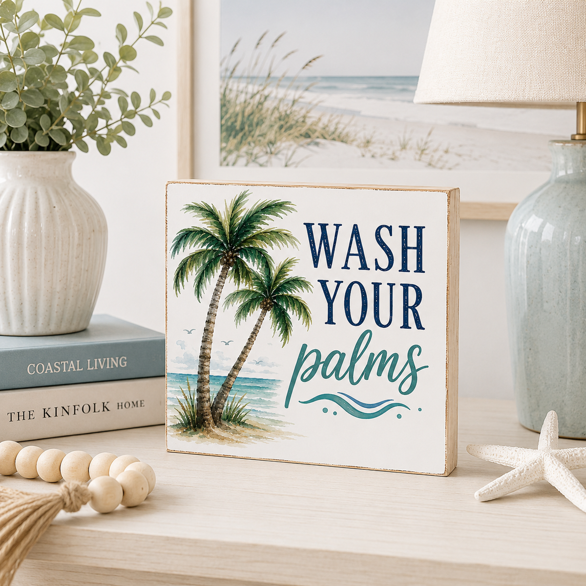 A rectangular decorative sign with a beach scene and the phrase “WASH YOUR palms” in navy and teal lettering, styled on a tabletop with a vase, books, lamp, beads, and a starfish.