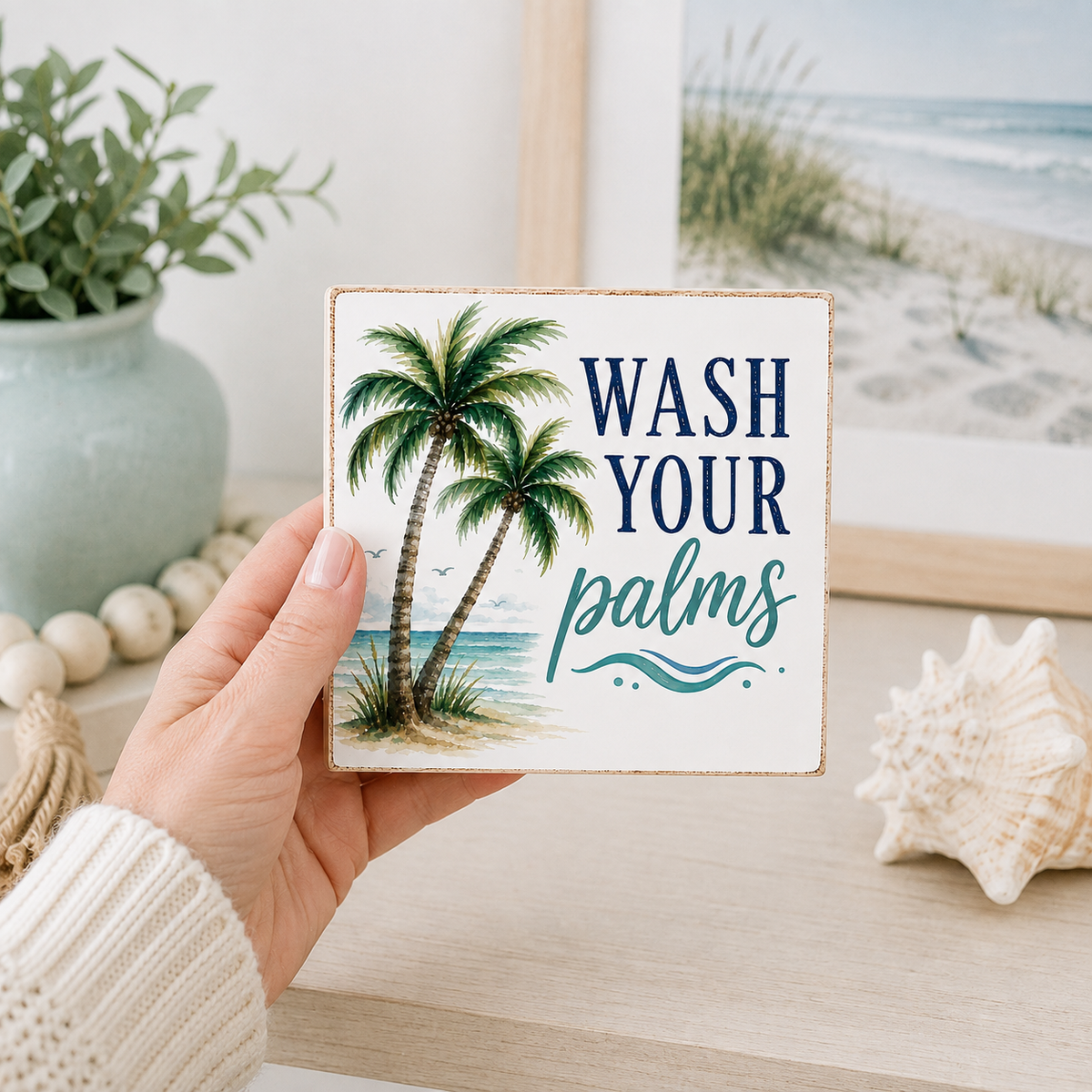 A hand holds a square decorative sign with a beach scene and the text 'WASH YOUR palms' in navy and teal lettering. The sign is shown in a styled home setting with a framed beach print, a seashell, and a potted plant in the background.
