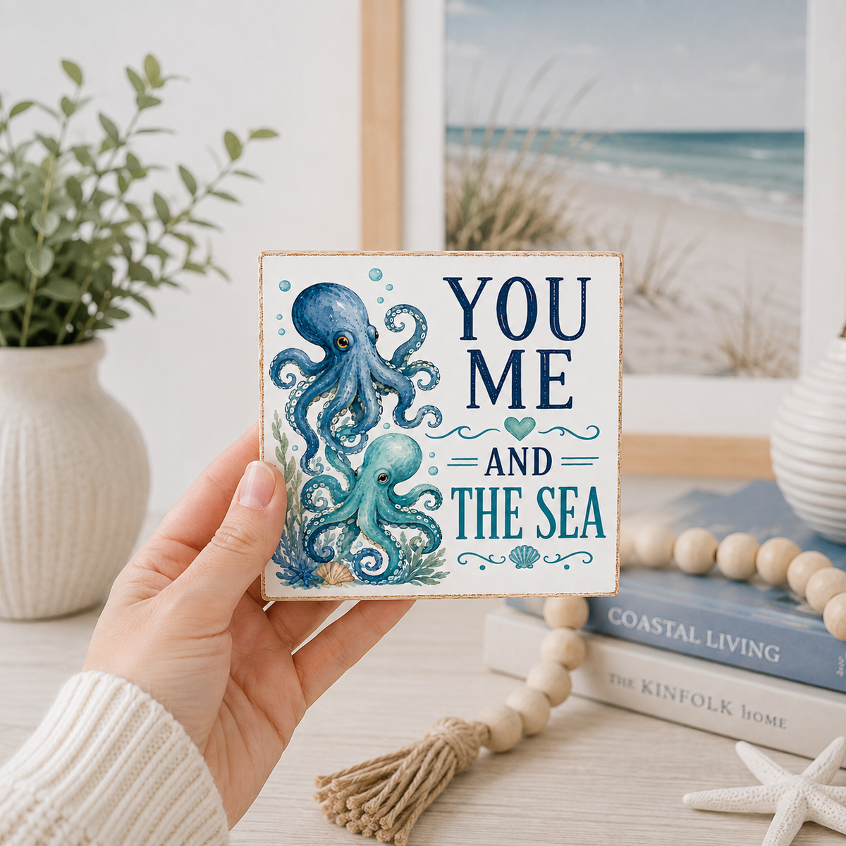 A hand holds a square decorative sign featuring a blue octopus and the text 'YOU ME AND THE SEA' in navy and teal lettering. The background shows a softly blurred beach scene, a potted plant, books, and a wooden bead garland.