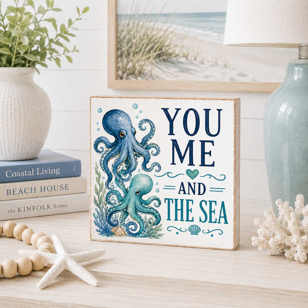 A square decorative sign with a nautical design featuring two blue octopuses and the text 'YOU ME AND THE SEA' in blue and teal lettering. The sign is styled on a light wood surface next to a white starfish, wooden beads, a vase with green leaves, books, a blue ceramic lamp, and a framed picture of a beach.