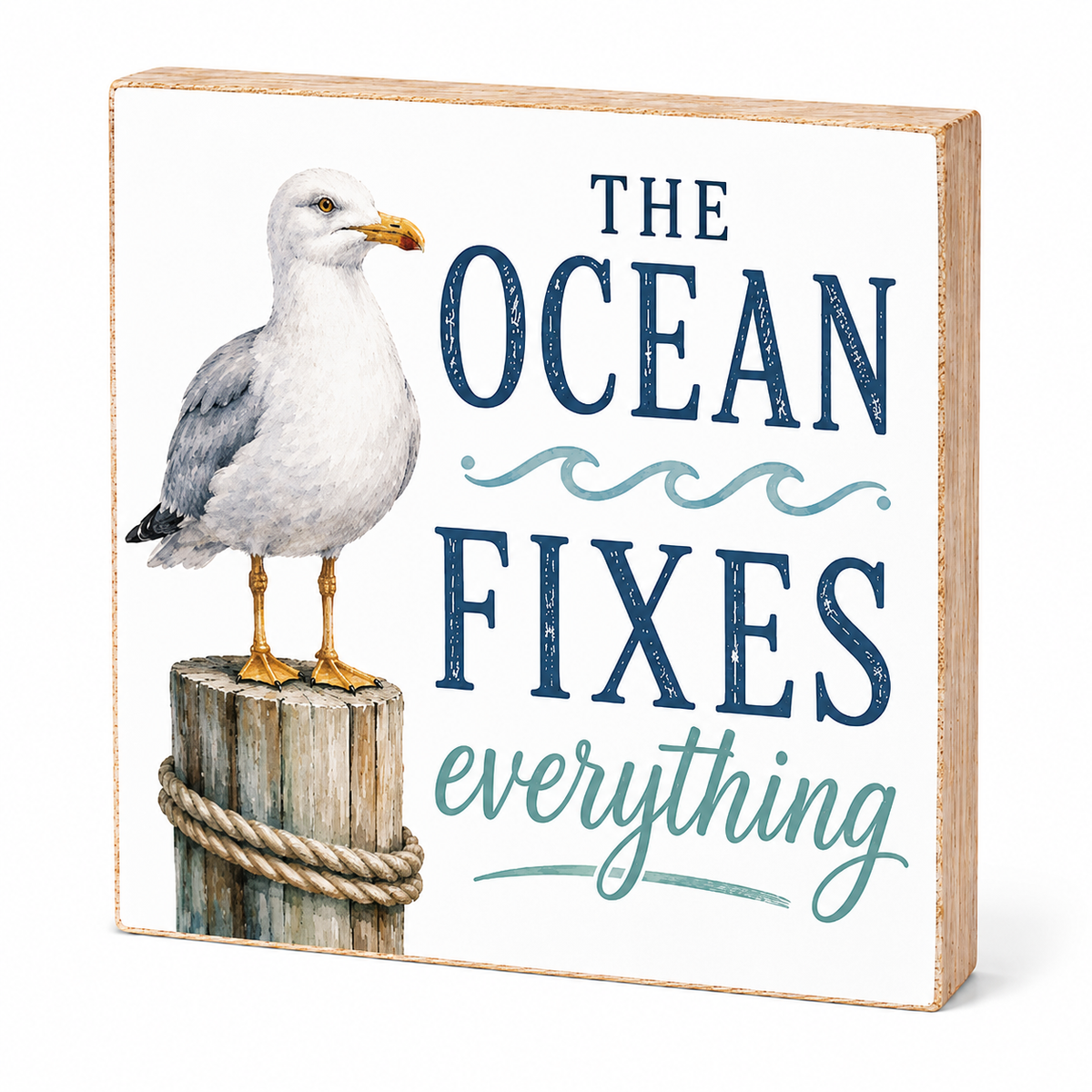 Square decorative wall sign with a seagull standing on a rope-bound post, next to the phrase “THE OCEAN FIXES everything” in navy and teal lettering on a white background, with a light wood edge.