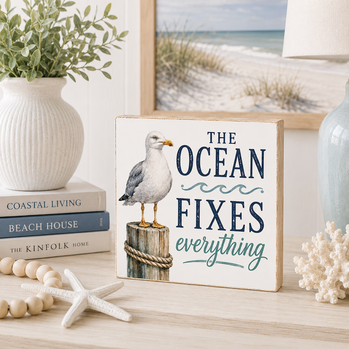 A decorative square sign with a seagull perched on a post and the phrase “THE OCEAN FIXES everything,” styled on a tabletop with books, a vase, beads, a starfish, and a beach scene in the background.