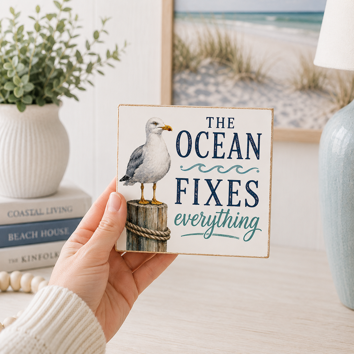 A hand holds a square decorative sign with a seagull standing on a post and the phrase “THE OCEAN FIXES everything” in navy and teal lettering. The background shows a softly blurred beach-themed interior with a vase, books, and a lamp.