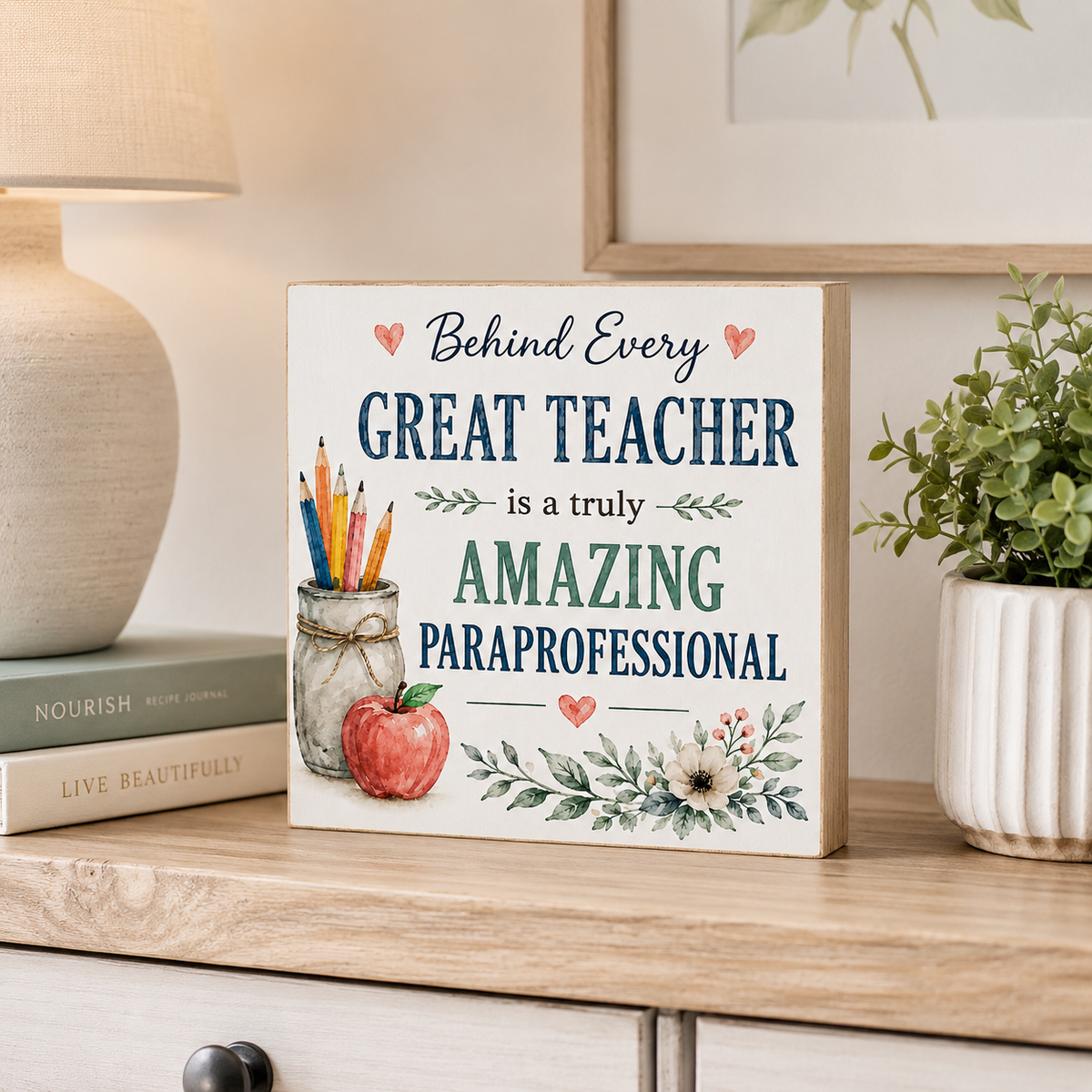 A decorative wooden sign with the phrase 'Behind Every GREAT TEACHER is a truly AMAZING PARaprofessional' in navy and green lettering, styled on a light wood dresser next to a lamp, books, and a potted plant.