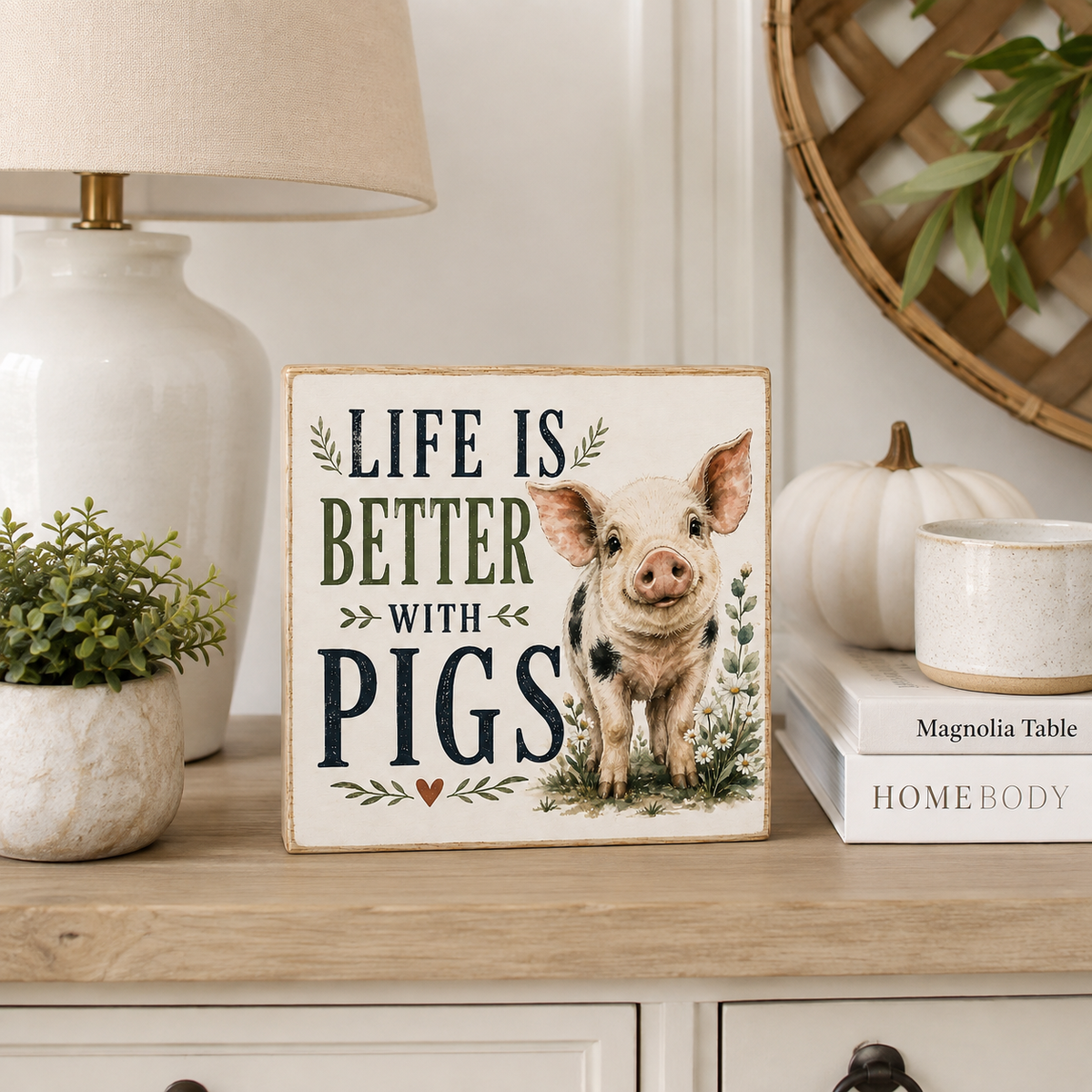 A square decorative sign with a pig illustration and the phrase “LIFE IS BETTER WITH PIGS” displayed on a wooden cabinet in a styled home interior setting.