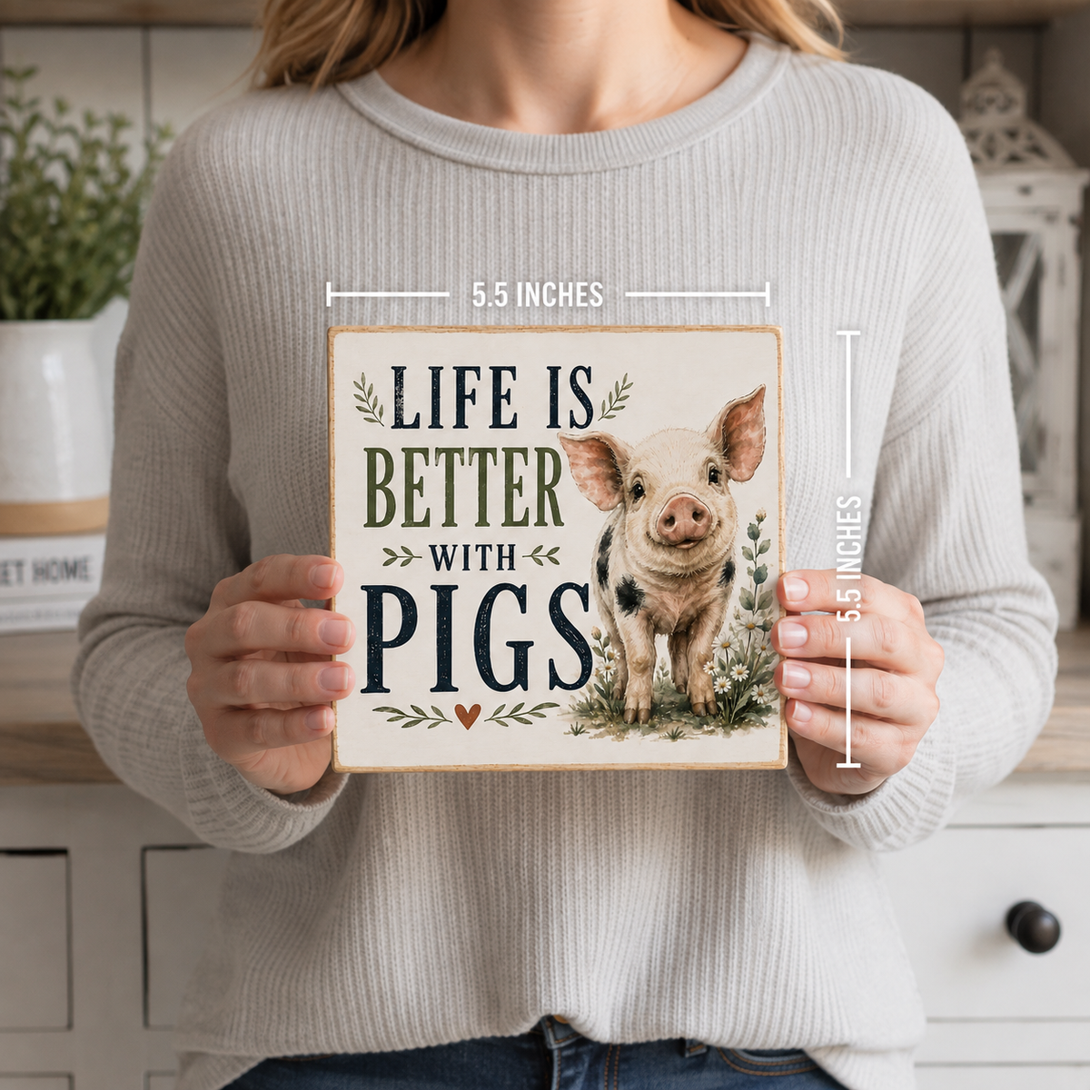 A woman in a ribbed light gray sweater holds a square decorative sign featuring the phrase “LIFE IS BETTER WITH PIGS” and an illustration of a pig. White measurement lines and text indicate the sign is 5.