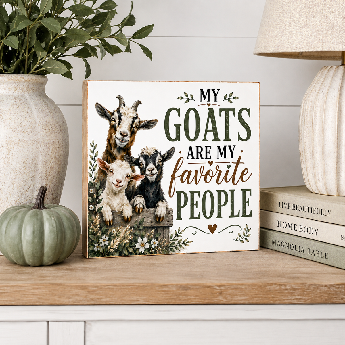 A decorative square sign with a rustic farm-style design featuring three goats and the text 'MY GOATS ARE MY favorite PEOPLE.' The sign is styled on a wooden shelf beside a vase with greenery, a small pumpkin, books, and a lamp in a home decor setting.
