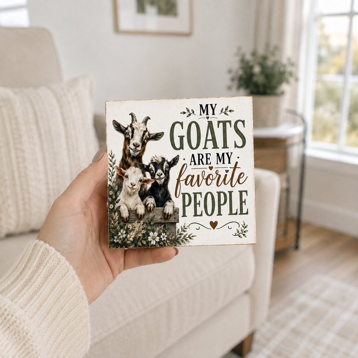 A hand wearing a cream knit sweater holds a square decorative sign with a family of goats and the text 'MY GOATS ARE MY favorite PEOPLE' in a cozy living room setting.