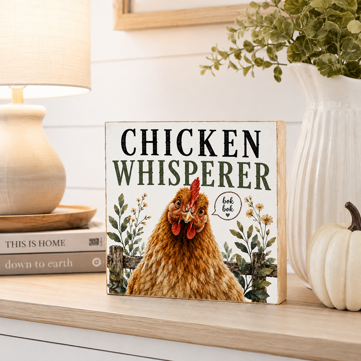 A decorative square wooden sign featuring the words “CHICKEN WHISPERER” and an illustration of a brown hen with a speech bubble reading “look book,” styled on a tabletop beside a lamp, books, a white pumpkin, and a white vase with greenery.