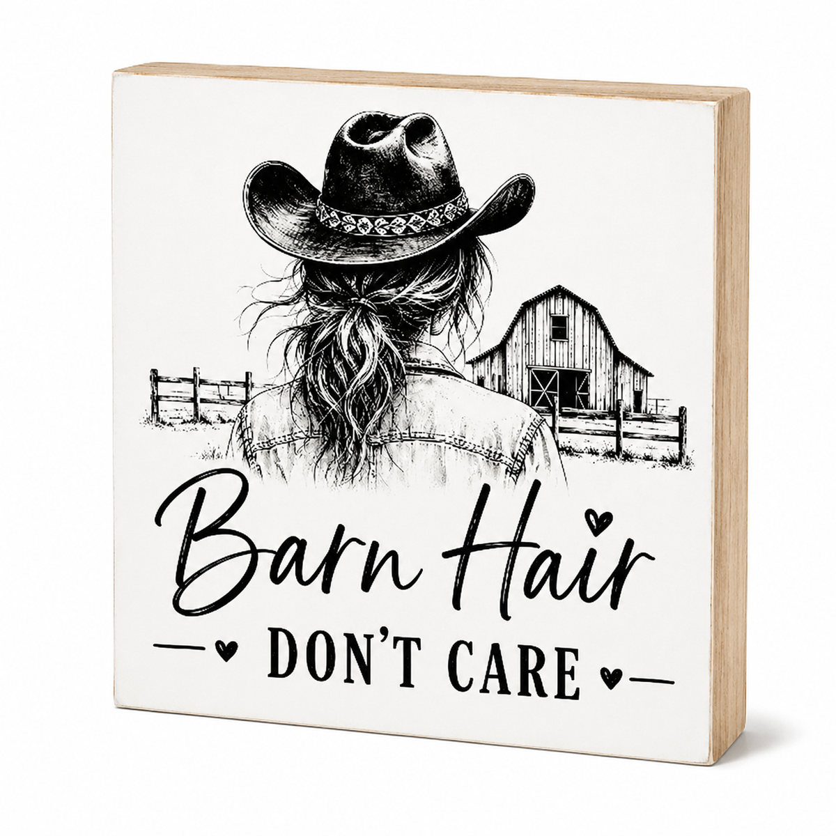 A square wooden sign with a white face and natural wood edges, featuring a black ink-style illustration of a woman from behind wearing a cowboy hat, with a barn and fence in the background and the text 'Barn Hair DON'T CARE' on the front.