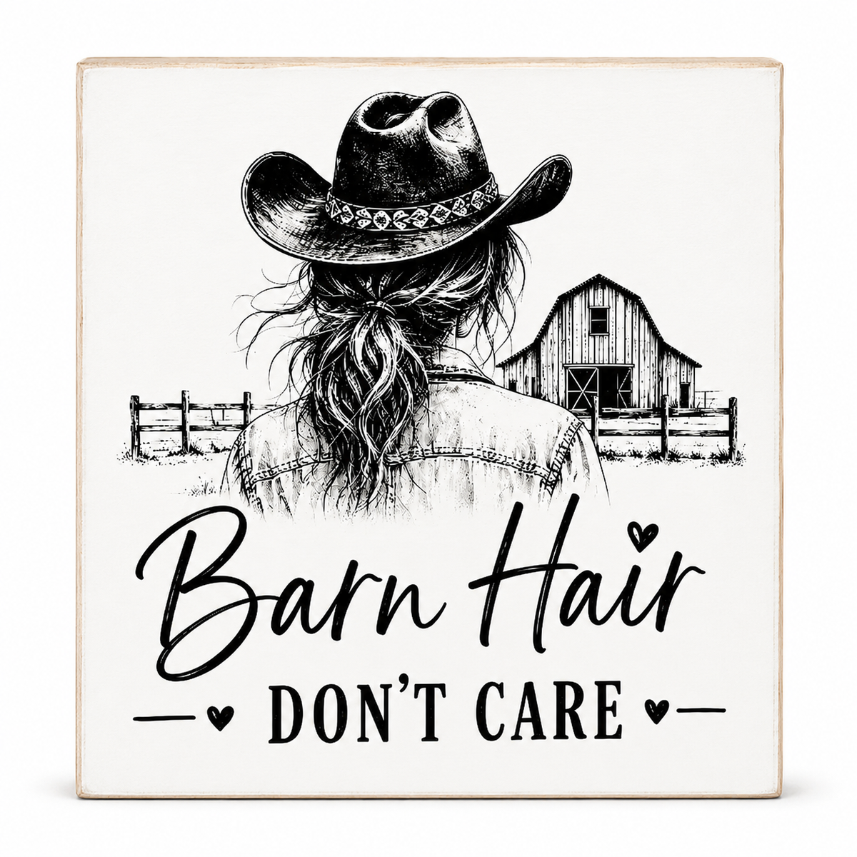 Square decorative sign with a black-and-white western illustration showing a woman from behind wearing a cowboy hat, with a barn and fence in the background and the text 'Barn Hair DON'T CARE' on an off-white surface with a light wood edge.
