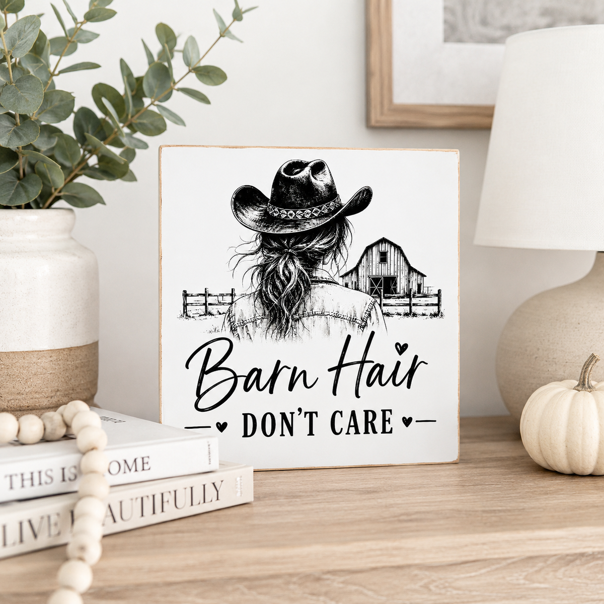 A square white decorative sign with a black illustration of a woman in a cowboy hat facing a barn, with the text 'Barn Hair DON'T CARE' on the front. The sign is styled on a wooden table with books, a ceramic vase with greenery, a lamp, and a small pumpkin.