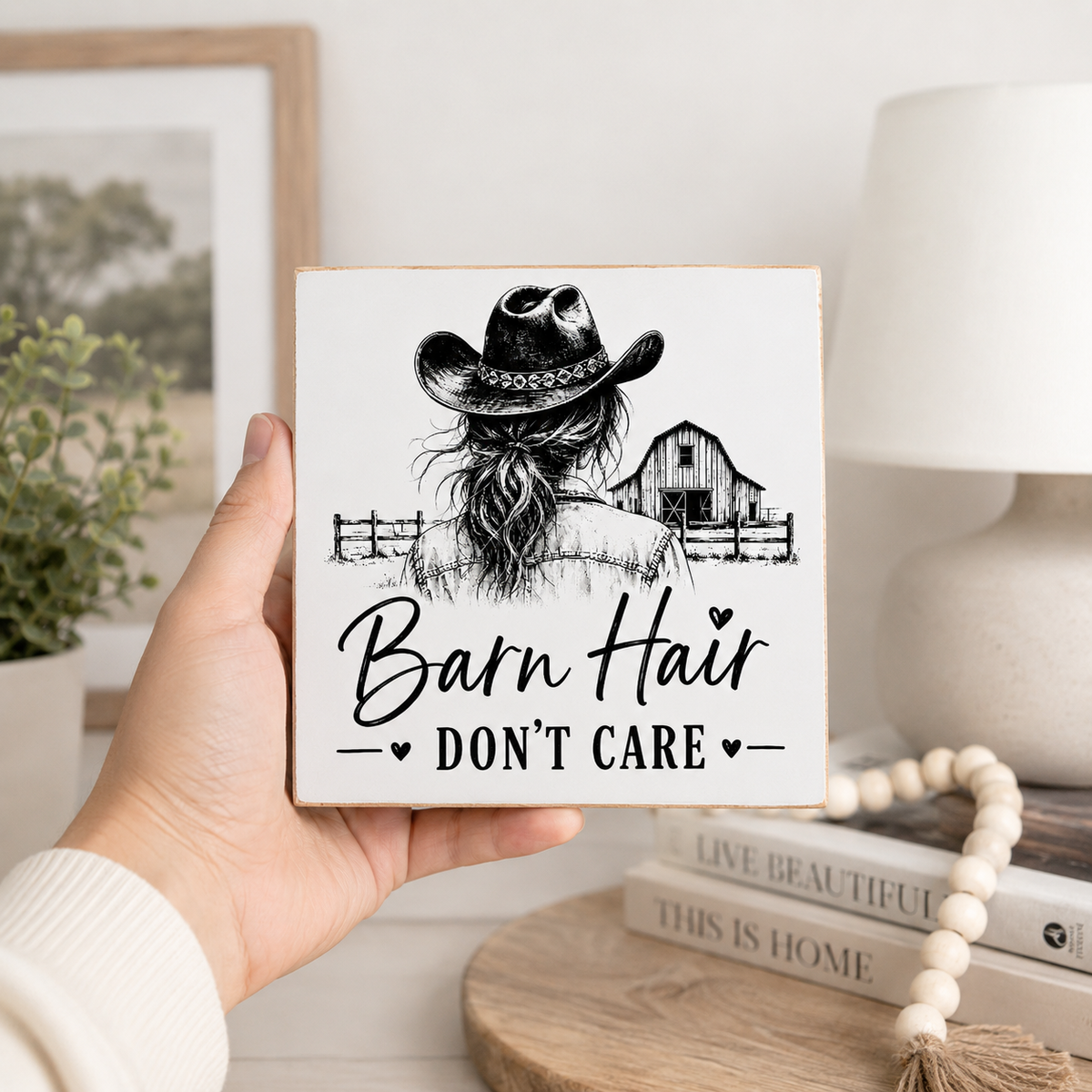 A hand holds a square white decorative sign with a black illustration of a woman in a cowboy hat and the text 'Barn Hair DON'T CARE'. The background shows a softly blurred home interior with a lamp, books, and a plant.