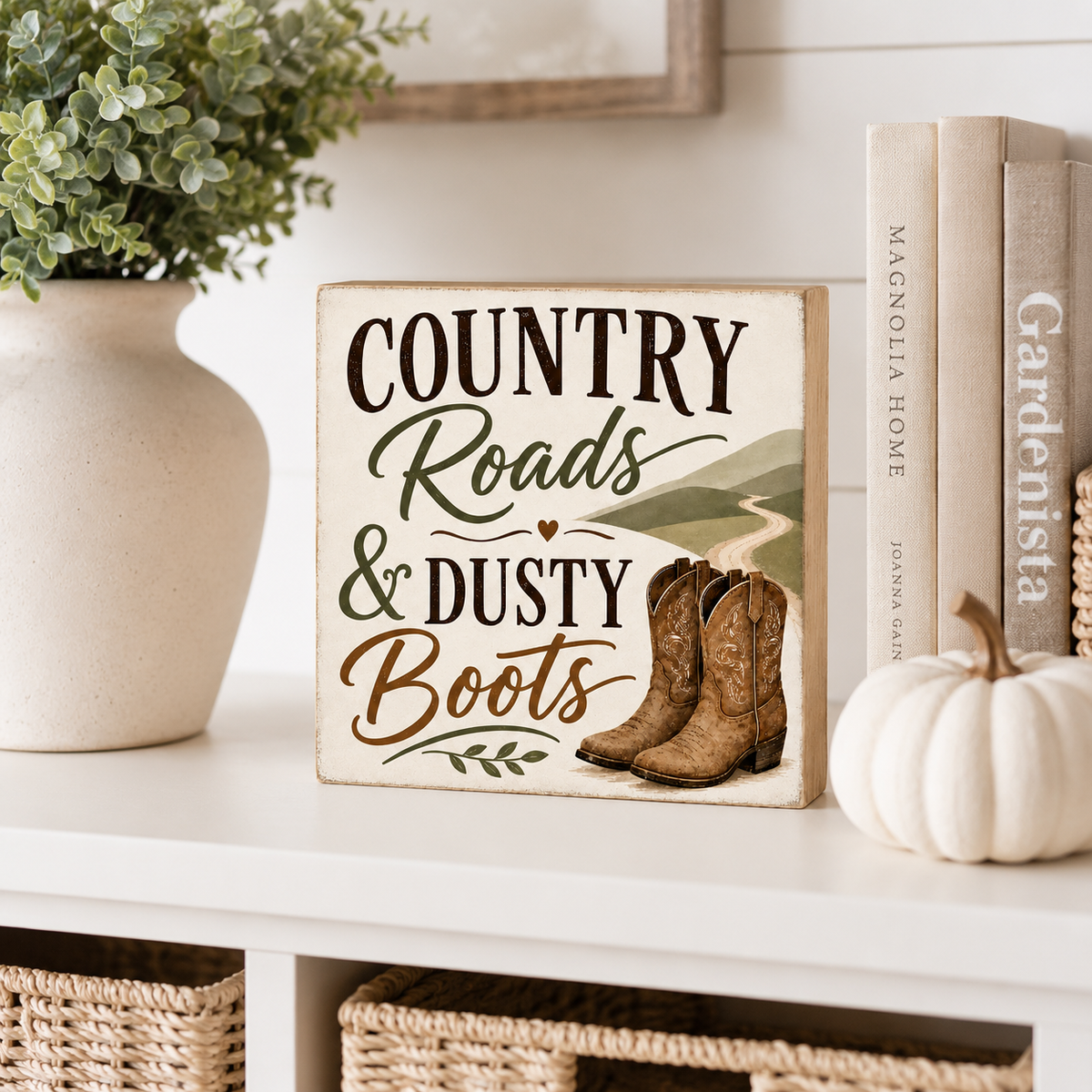 A square decorative sign reading “COUNTRY Roads & DUSTY Boots” sits on a white shelf beside a white vase with greenery, a small white pumpkin, and books. The sign has a distressed cream background with dark brown lettering, green script text, a small red heart, a green landscape illustration, and a pair of brown cowboy boots.