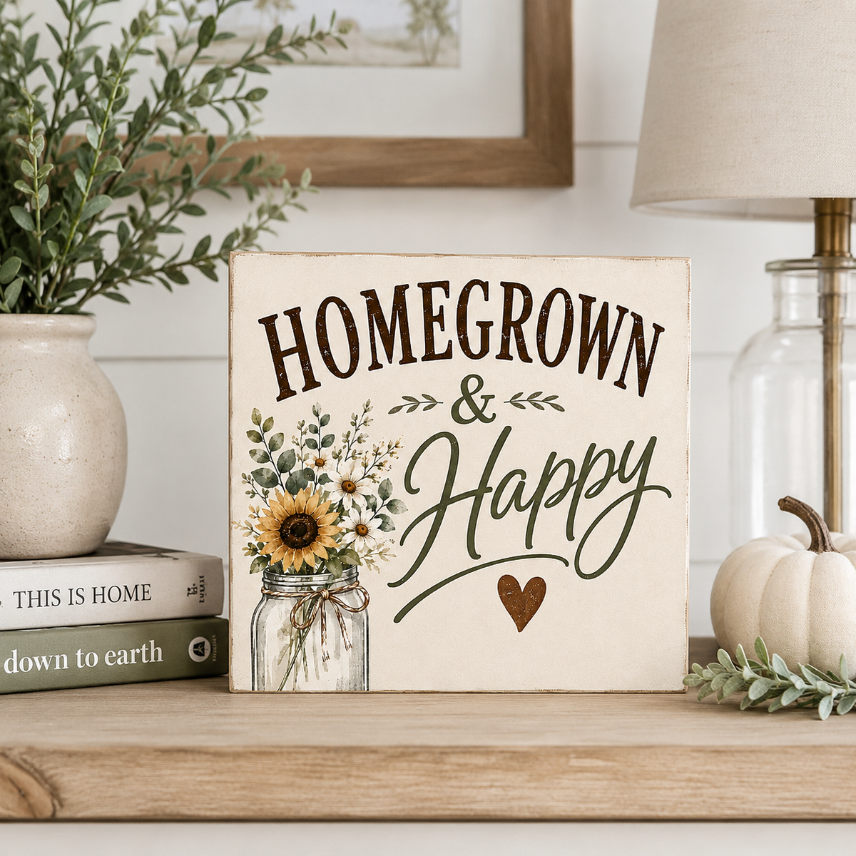 A square decorative sign reading “HOMEGROWN & Happy” with a mason jar flower illustration and small heart, displayed on a wooden shelf in a styled home setting with books, a plant, a lamp, and a small pumpkin.