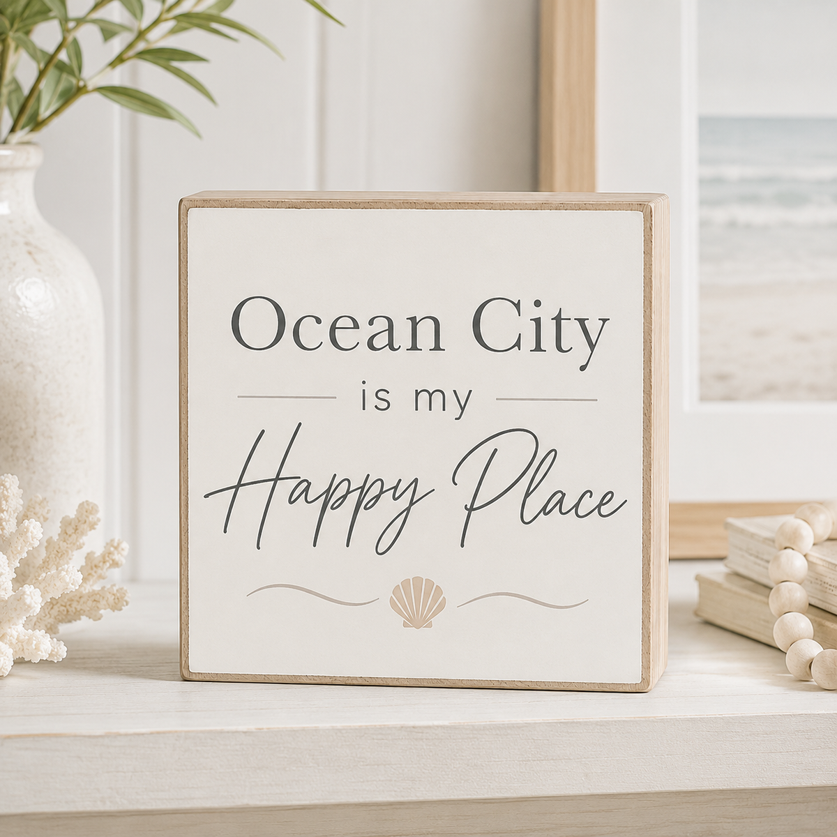 A square decorative wooden sign with a white face and natural wood border, reading “Ocean City is my Happy Place” with a small shell motif, styled on a tabletop with a vase, framed beach photo, and beads in the background.