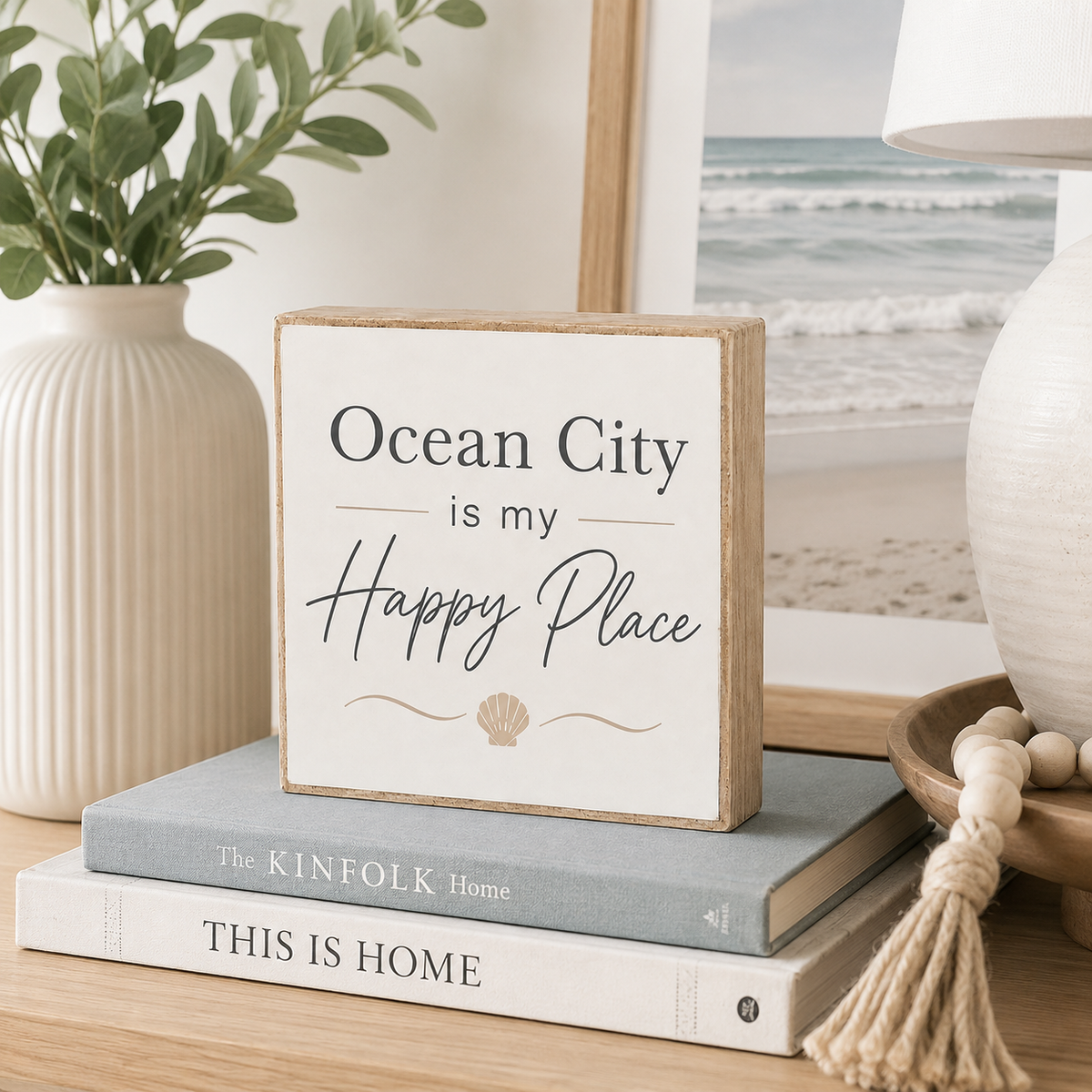 A square decorative wooden sign with the phrase 'Ocean City is my Happy Place' and a small seashell motif, styled on top of two books in a coastal-themed room setting.