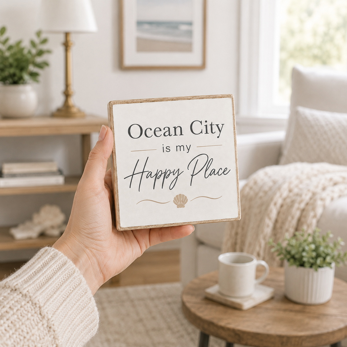 A hand holds a square wooden sign that reads 'Ocean City is my Happy Place' with a small seashell graphic, set against a cozy, softly blurred living room background.