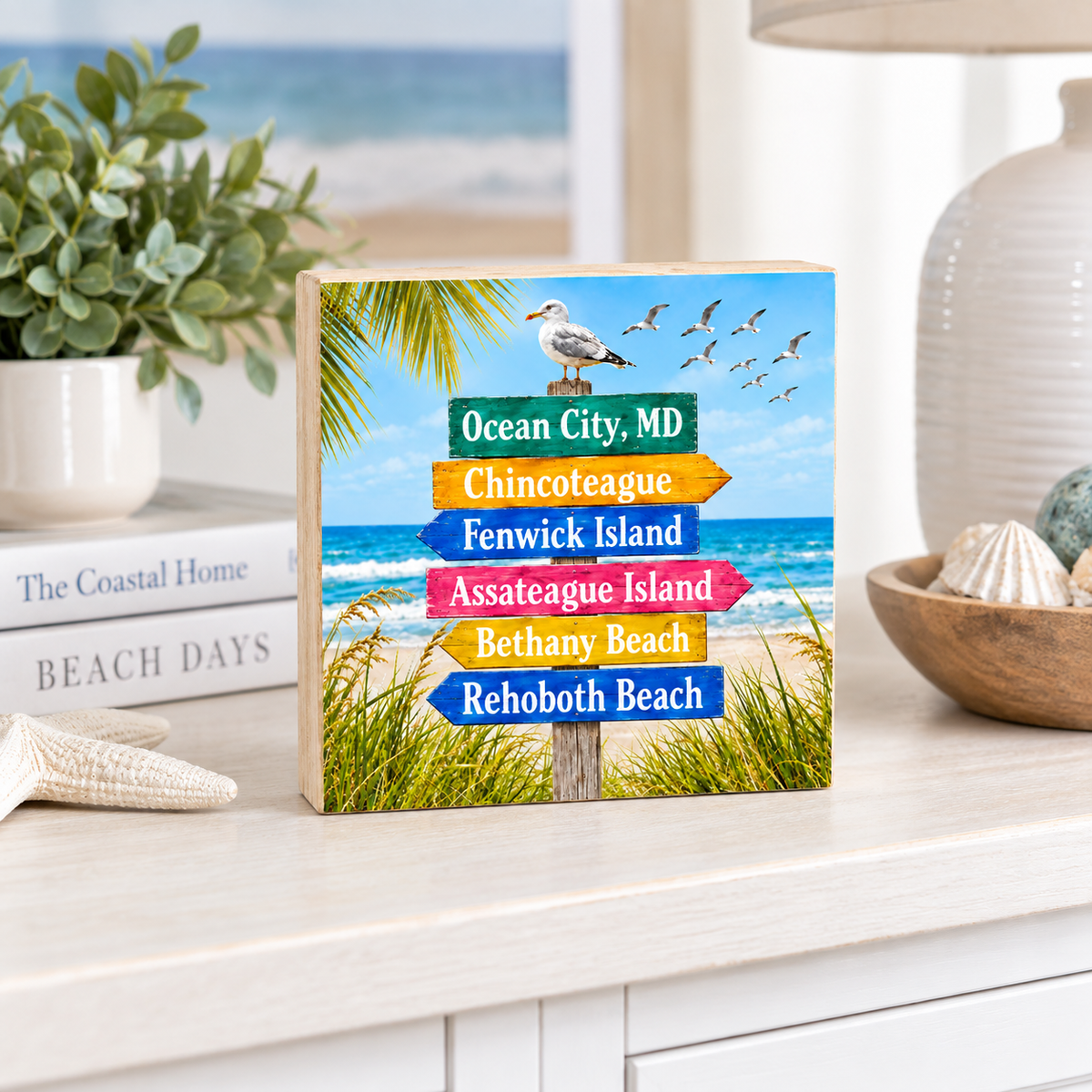 A personalized wooden sign with a beach scene background and colorful directional arrow signs reading Ocean City, MD, Chincoteague, Fenwick Island, Assateague Island, Bethany Beach, and Rehoboth Beach, displayed on a light wood console table in a coastal-themed room setting.