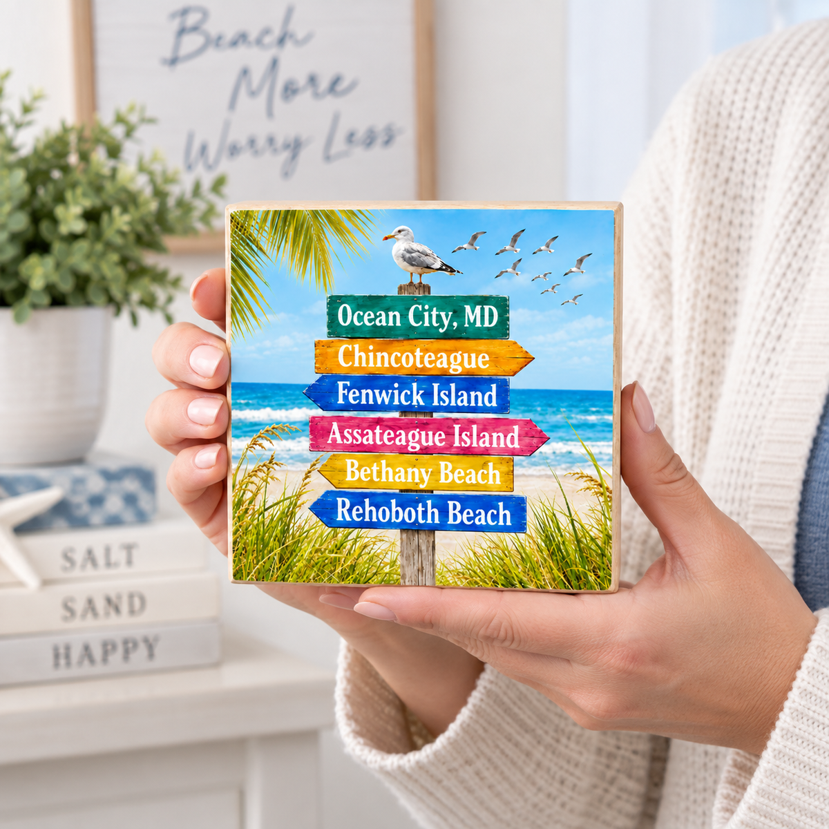 A person in a cream knit sweater holds a square wooden sign featuring a beach scene with a seagull and colorful directional signs reading Ocean City, MD; Chincoteague; Fenwick Island; Assateague Island; Bethany Beach; Rehoboth Beach.