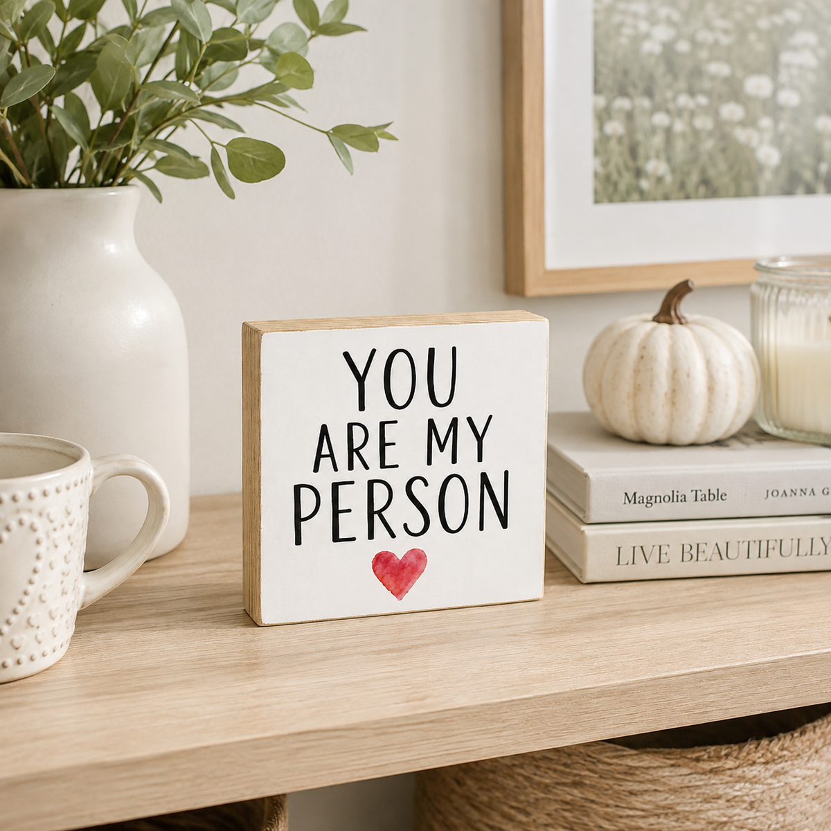 A square wooden sign with a white face and natural wood edges reads 'YOU ARE MY PERSON' in black lettering with a small red heart below. The sign sits on a light wood shelf styled with a white mug, a white vase with greenery, books, a small pumpkin, and a candle.