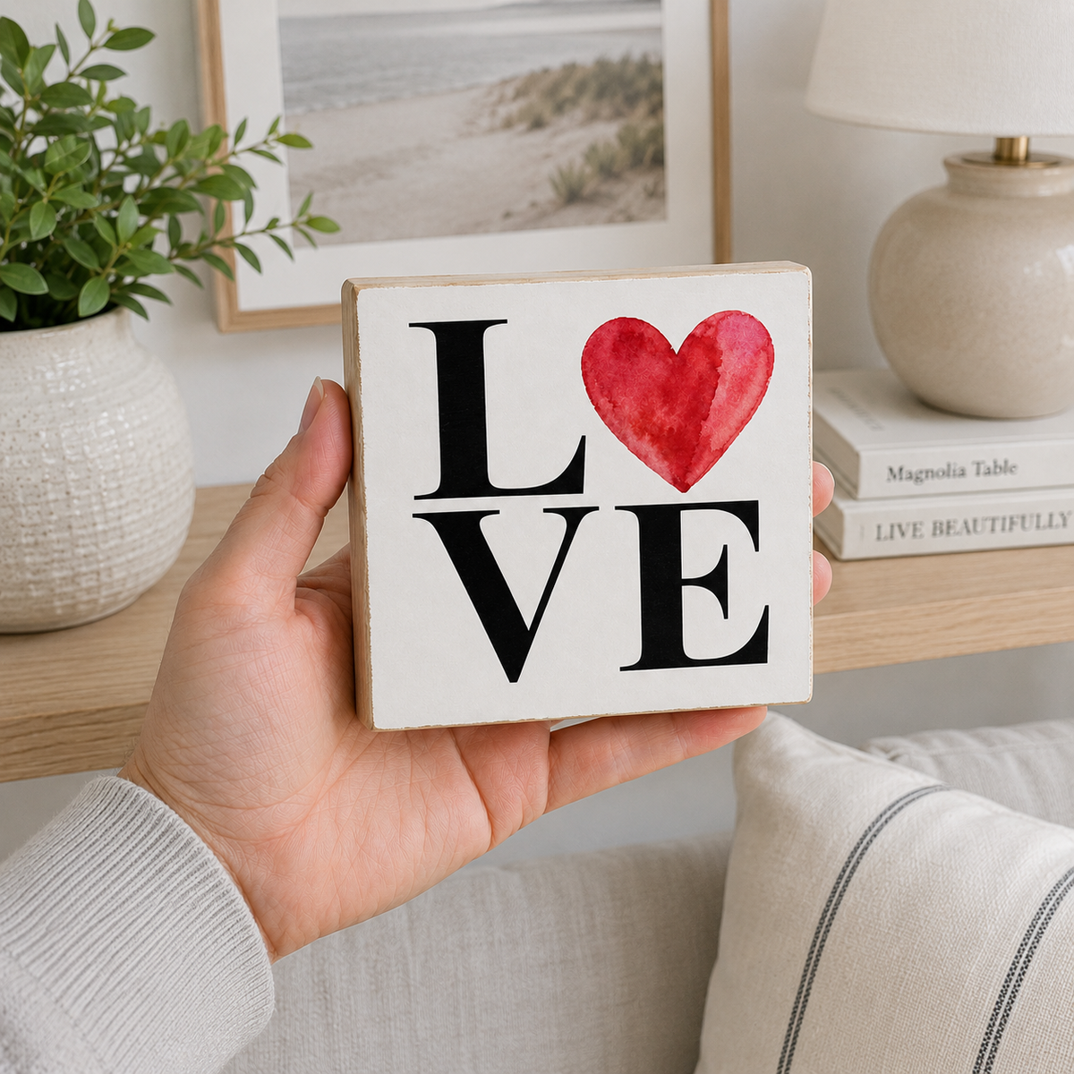 A hand holds a square white wooden sign with the word LOVE, where the O is replaced by a red watercolor heart. The background shows a cozy home interior with a shelf, framed picture, lamp, and plant.