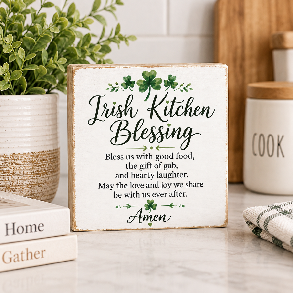 A square decorative sign with the words 'Irish Kitchen Blessing' and a short prayer, styled on a kitchen counter with a plant, books, and a canister in the background.