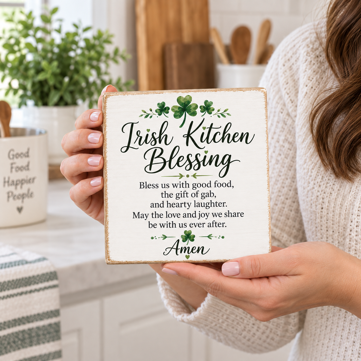 A person in a white knit sweater holds a square decorative sign that reads “Irish Kitchen Blessing” with a St. Patrick’s Day prayer and shamrock accents.