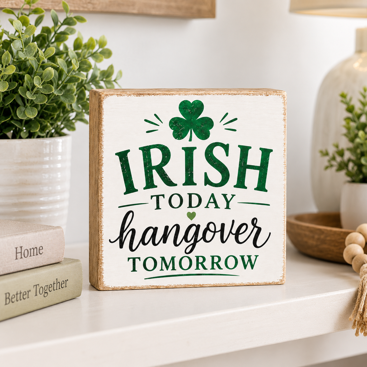 A rectangular wooden sign with a distressed white face and natural wood edges, printed with the phrase “IRISH TODAY hangover TOMORROW” in green and black lettering beneath a green shamrock. The sign is styled on a white shelf next to potted green plants and decorative books.