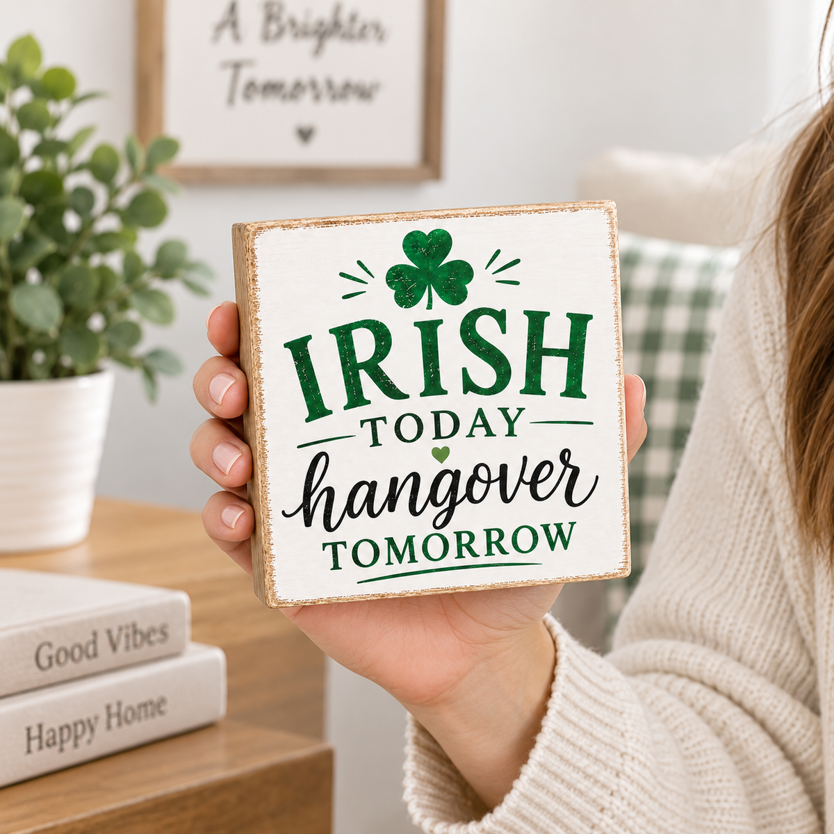 A person in a cream sweater holds a square wooden sign that reads 'IRISH TODAY hangover TOMORROW' with a green shamrock and dark green text. The background shows a cozy room with a potted plant, framed wall art, and books on a wooden table.