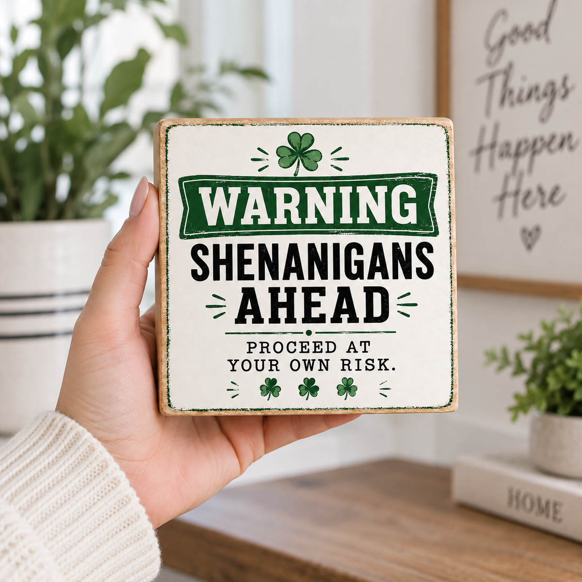A hand holds a square decorative sign with a rustic wood border and a white face printed with the phrase “WARNING SHENANIGANS AHEAD” in black text, green shamrocks, and a green warning banner. The background shows a softly blurred home interior with plants and a framed sign.