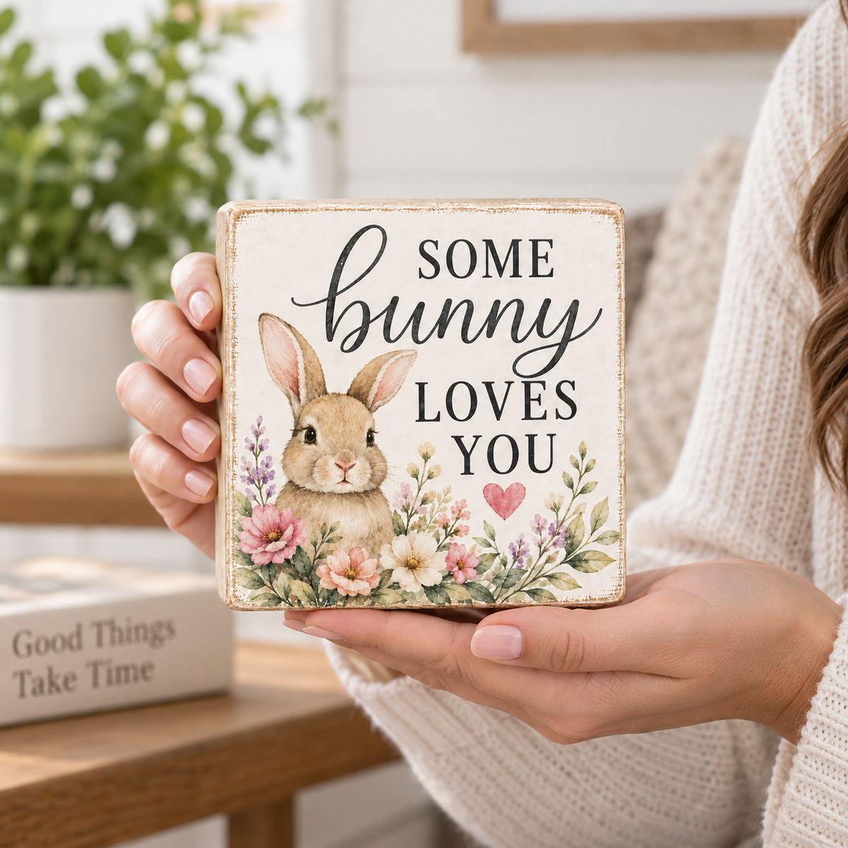 A person in a cream knit sweater holds a square decorative wooden sign that reads “Some bunny loves you” with a watercolor-style bunny and floral border. The background shows a softly blurred indoor setting with a plant and table.