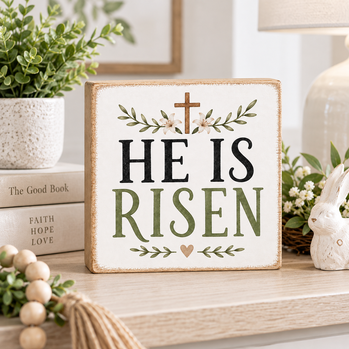 A square decorative wooden sign with the words “HE IS RISEN” in black and muted green lettering, topped with a small cross and floral wreath. The sign is styled on a tabletop in a bright home setting with a plant, books, a lamp, and a small white rabbit figurine nearby.