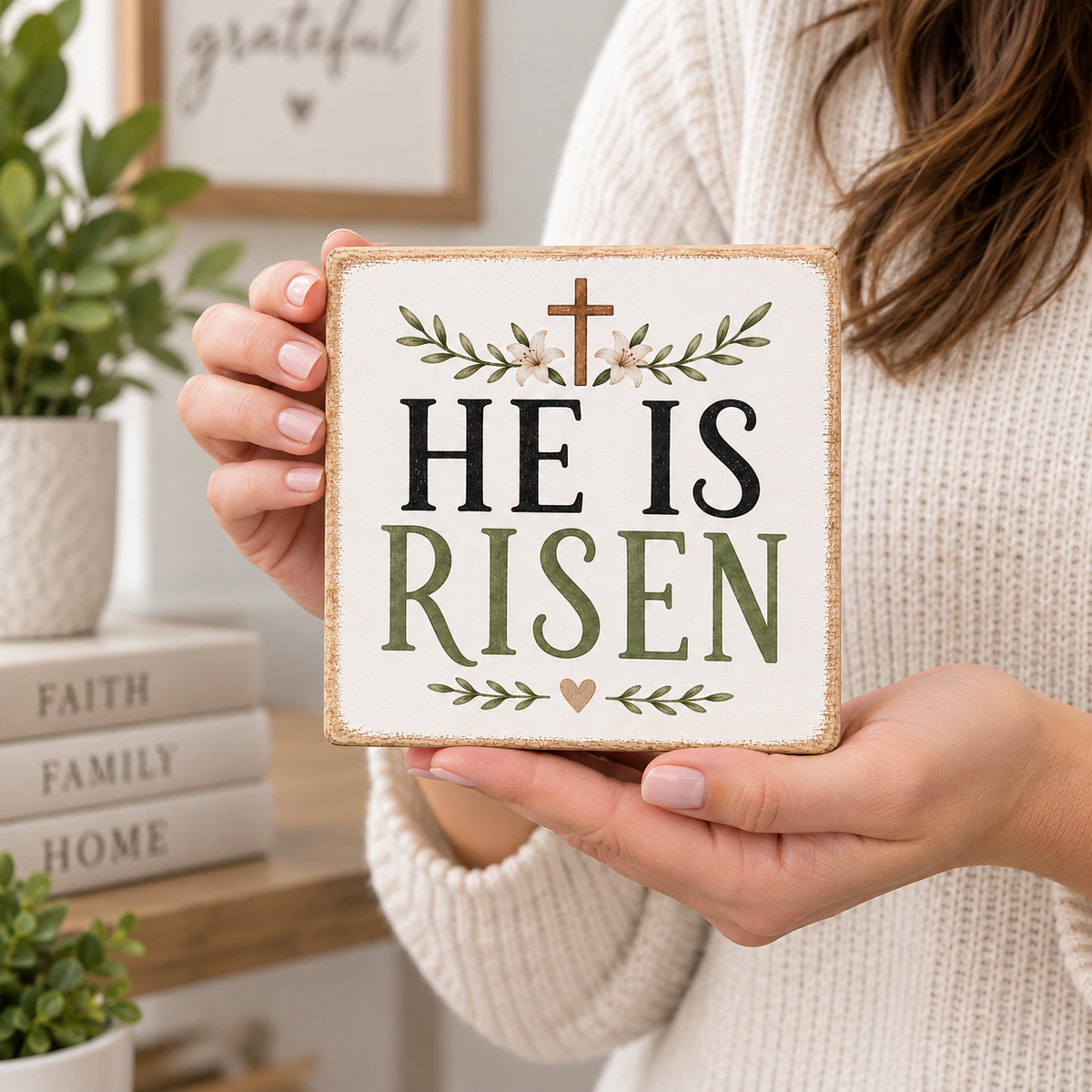 A person in a cream knit sweater holds a square decorative sign that reads “HE IS RISEN” with a small cross, floral vine, and heart details. The scene is a softly lit indoor setting with books and greenery in the background.