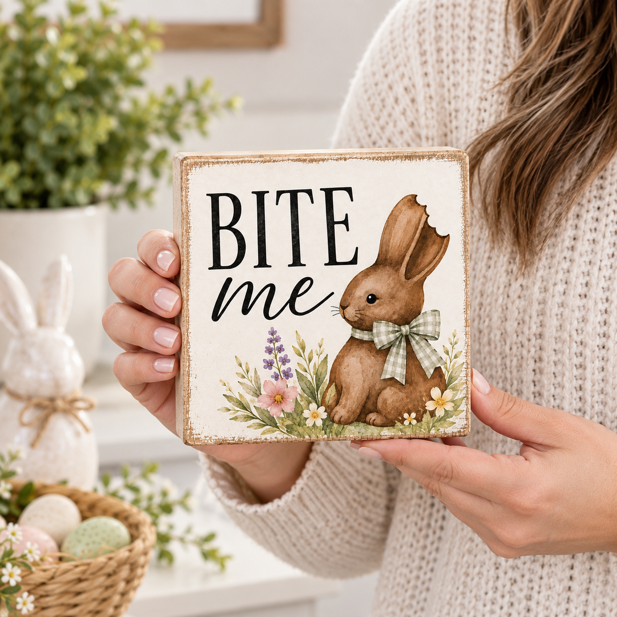 A person in a cream knit sweater holds a square decorative sign with a rustic bunny illustration and the words “BITE me” in black script. The sign has a warm off-white background with a brown rabbit, green bow, and small floral details.