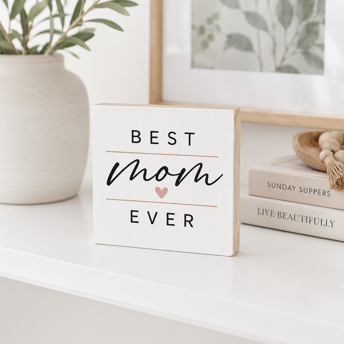 A square white wooden sign with the words 'BEST mom EVER' in black lettering and a small pink heart, displayed on a white shelf next to books and a plant.