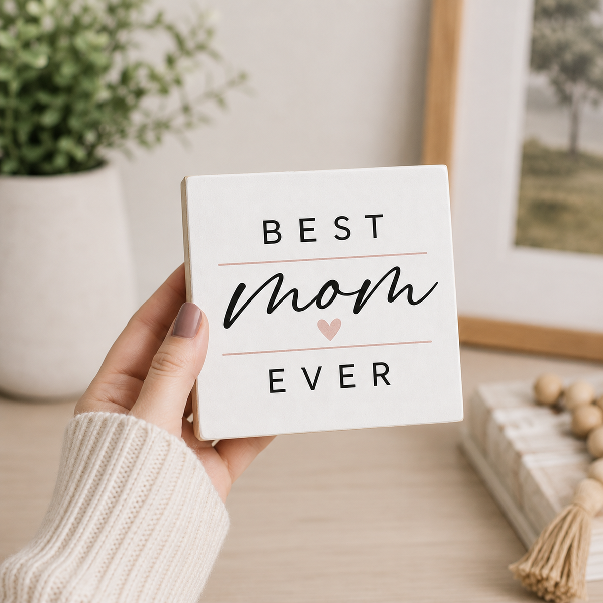 A hand wearing a cream knit sweater holds a square white ceramic sign that reads 'BEST mom EVER' with a small pink heart. The background shows a softly blurred home interior with a potted plant, framed picture, and decorative items on a tabletop.