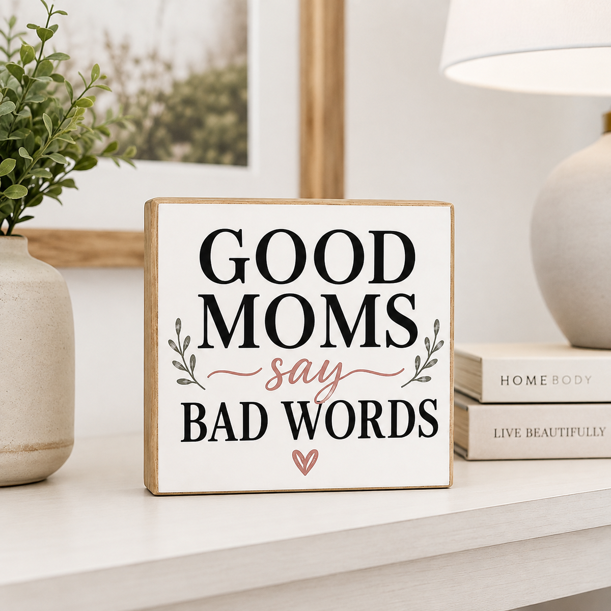 A square decorative sign with the phrase “GOOD MOMS say BAD WORDS” in black and dusty pink lettering, displayed on a light tabletop in a styled home setting with a plant, lamp, and books.