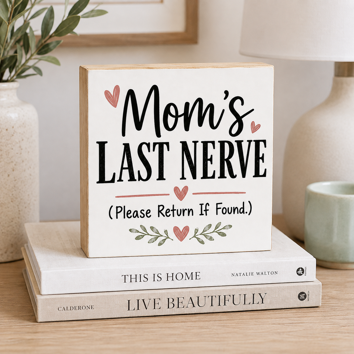 A square wooden sign with the phrase 'Mom's Last Nerve (Please Return If Found.)' in black lettering with small pink hearts and leaf accents, displayed on top of two books in a softly styled home interior.