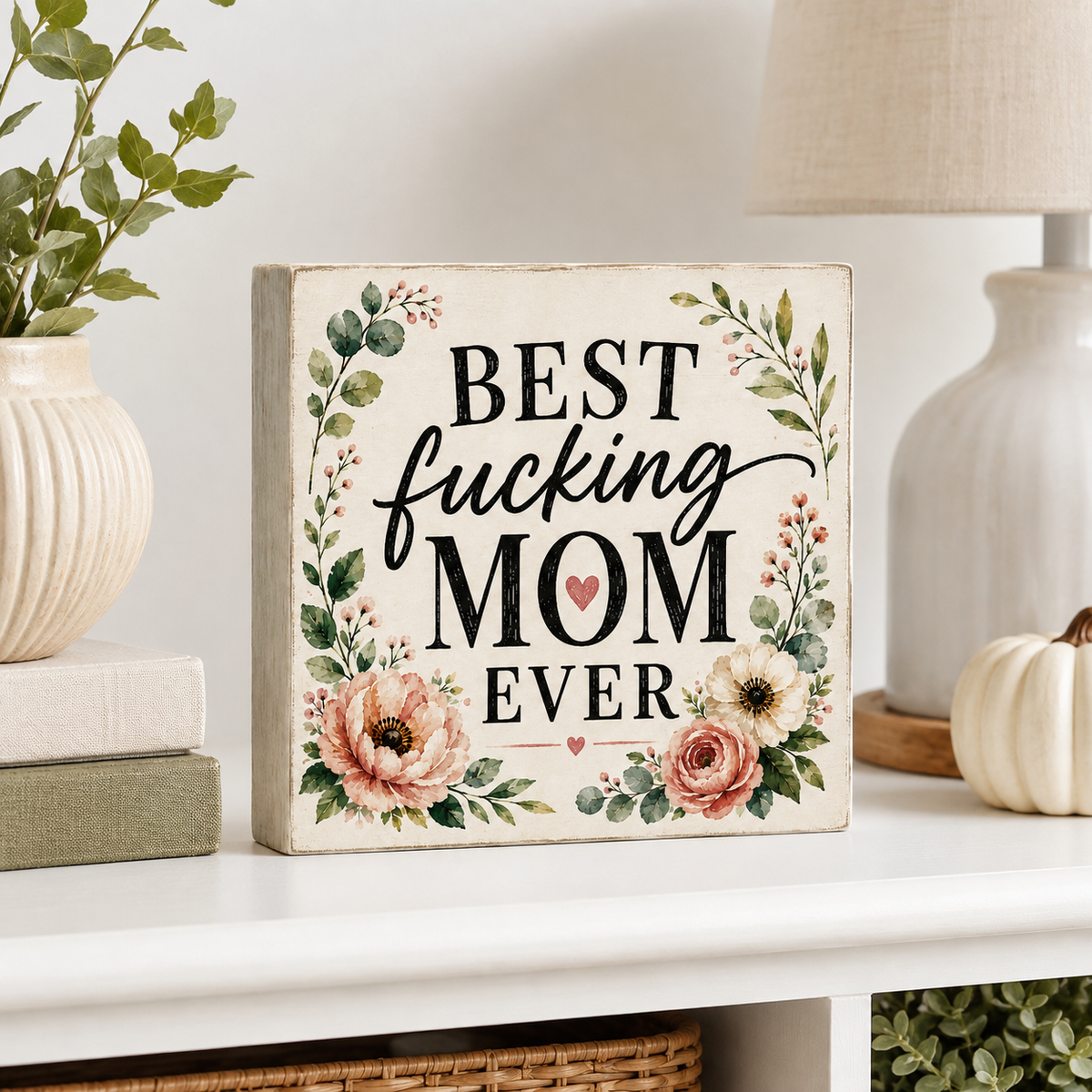 A rectangular decorative sign with the phrase “BEST fucking MOM EVER” in black lettering, surrounded by watercolor-style flowers and greenery, displayed on a white shelf in a styled home setting.