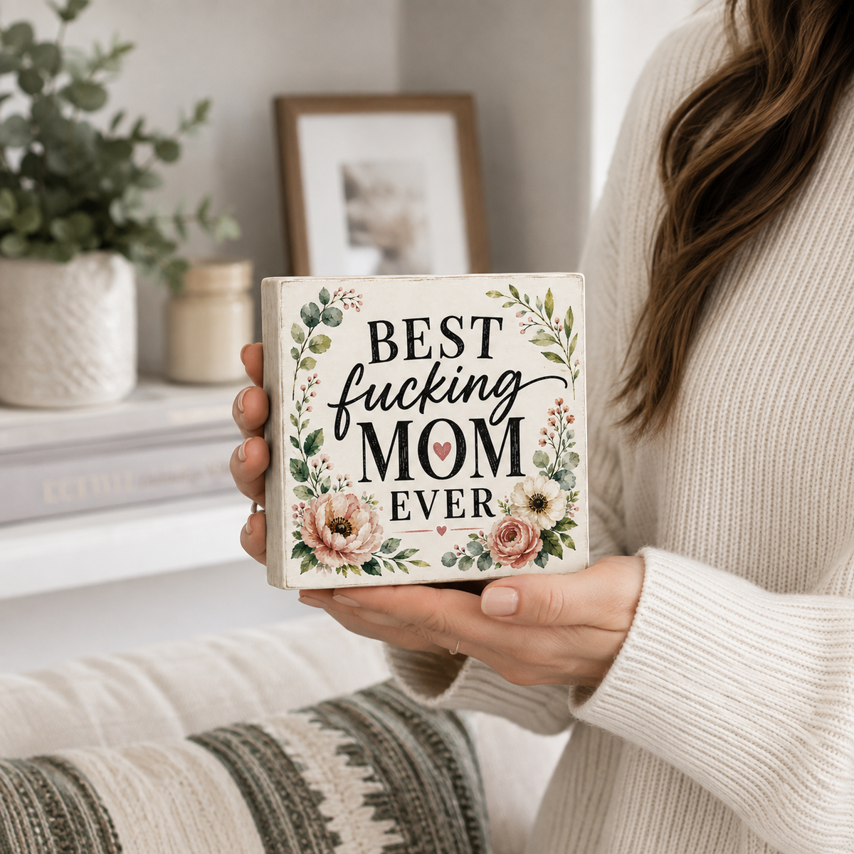 A person in a cream knit sweater holds a square decorative sign that reads “Best fucking MOM EVER” with floral artwork. The scene is a softly lit indoor setting with a plant, books, and framed art in the background.