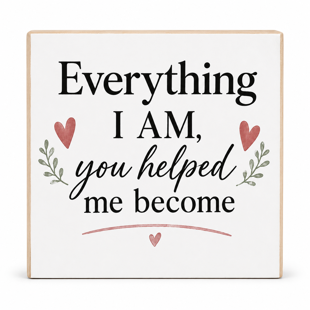 A square decorative wooden sign with a white face and natural wood edges, featuring the printed phrase 'Everything I AM, you helped me become' in black lettering, accented with pink hearts and small green leaf motifs.
