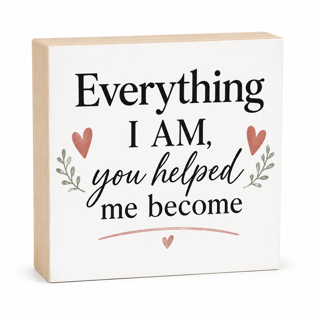 A square decorative wooden block sign with a white front panel and natural light wood sides. The front reads, 'Everything I AM, you helped me become' in black lettering, decorated with two pink hearts, small green leaf sprigs, and a pink curved line at the bottom.