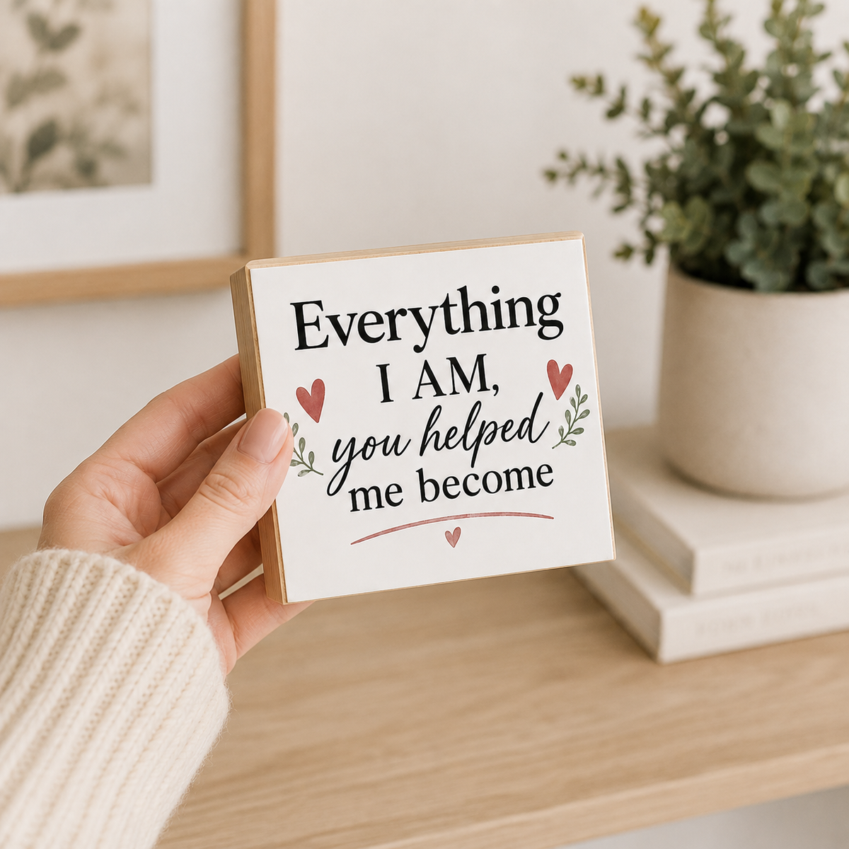 A hand wearing a cream knit sweater holds a square white sign with a kraft paper border. The sign features the text 'Everything I AM, you helped me become' in black script and serif fonts, decorated with small red hearts and green leafy sprigs.
